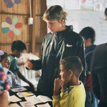 The image depicts a young man standing beside a table with children, surrounded by papers, in a classroom setting with wooden walls and colorful decorations.