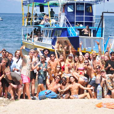 The image depicts a group of approximately 30 people posing for a photo on a sandy beach, with a large blue boat in the background and a body of water behind it.