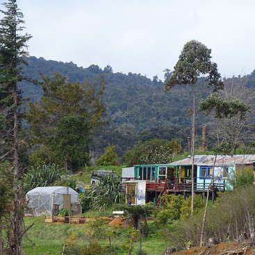 The image depicts a serene rural scene with a house, greenhouse, and car amidst lush trees and vegetation, set against a mountainous backdrop on a cloudy day.