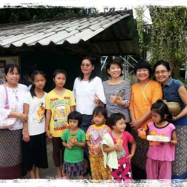 The image depicts a group of women and children standing together in front of a building with a tiled roof, posing for a photo.