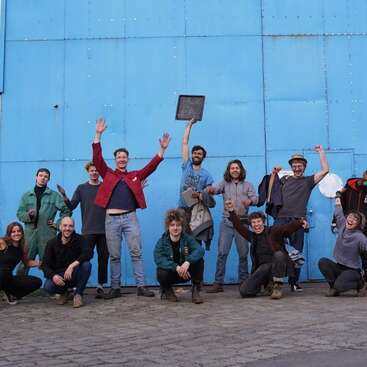 The image depicts a group of 13 people posing for a photo in front of a large blue wall, with some individuals raising their arms in celebration.