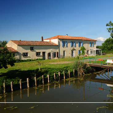 The image depicts a serene scene of a large, two-story house with a distinctive orange-tiled roof, situated beside a tranquil body of water and lush greenery.