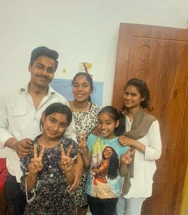 A imagem mostra uma família de cinco pessoas posando para uma foto, com o pai em pé atrás da mãe e das três filhas, todas fazendo sinais de paz.