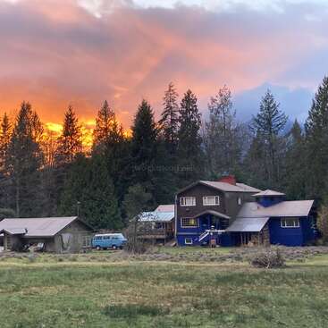 The image depicts a serene rural scene featuring a large house with multiple stories, a blue van, and a lush grassy field, set against a vibrant sunset sky.