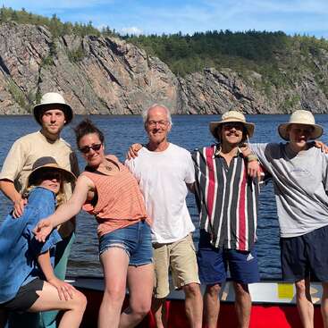 The image depicts a group of six people posing for a photo on a canoe in front of a serene lake and rocky cliffside, set against a bright blue sky.