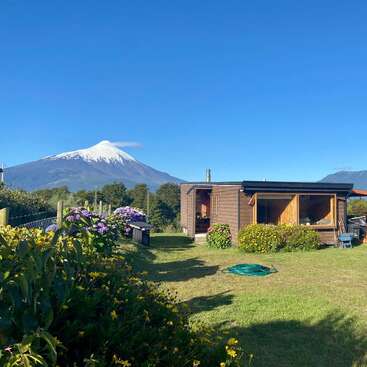 A cozy wooden cabin sits on a lush green lawn, surrounded by flowers, with a snow-capped volcano and mountains in the background under a clear sky.