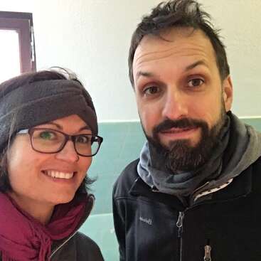 The image shows a selfie of a man and woman, both wearing winter clothing, standing in front of a white wall with green tiles at the bottom.
