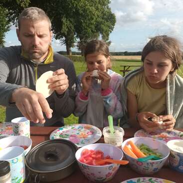 The image depicts a man and two girls sitting at a picnic table, enjoying a meal together, with the man preparing food on a plate.