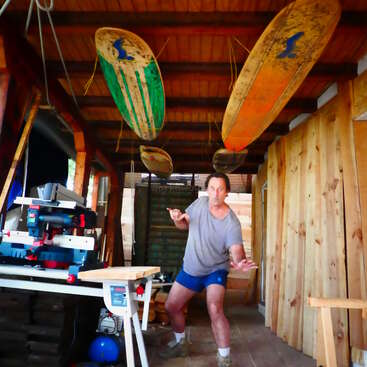 The image depicts a man standing in a workshop, surrounded by surfboards and woodworking tools, with a wooden ceiling and walls, conveying a sense of creativity and activity.
