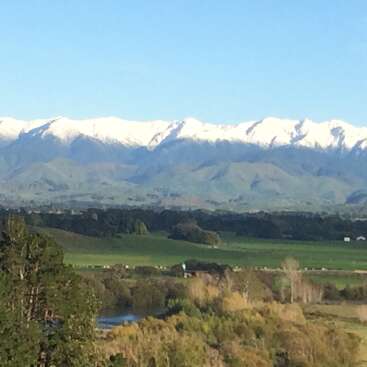 The image depicts a serene landscape featuring a snow-capped mountain range, lush green fields, and trees, set against a clear blue sky.