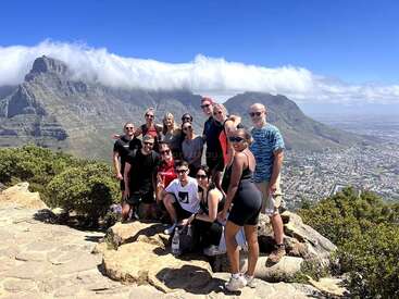The image depicts a group of 13 people posing on a rocky outcrop, with a cityscape and mountain range visible in the background.