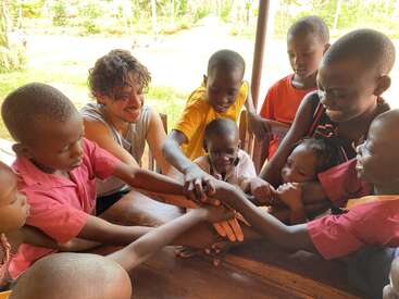 The image depicts a group of children and an adult sitting in a circle, holding hands, with a serene outdoor setting featuring trees and grass in the background.