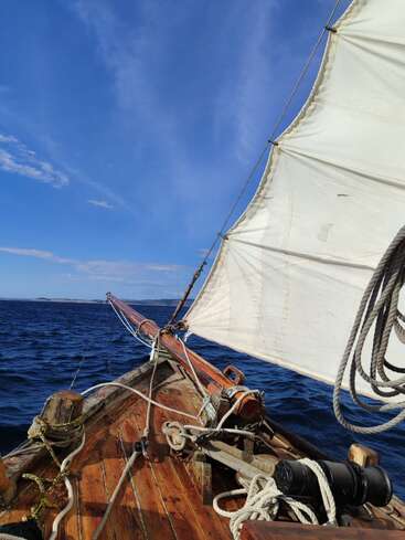 The image depicts a wooden boat with a white sail on a vast body of water, set against a blue sky with scattered clouds.