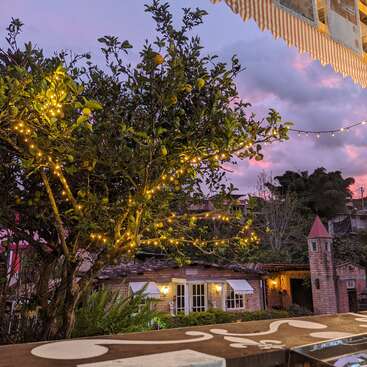 The image depicts a serene outdoor setting at dusk, featuring a tree adorned with string lights, a charming brick building, and a vibrant pink and purple sky.