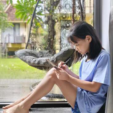 A young girl sits indoors by a glass window, focused on using her tablet with a stylus. Outside, a hanging chair and greenery are visible.