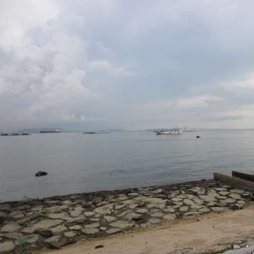 The image depicts a serene body of water with a stone wall and sandy beach in the foreground, under a cloudy sky with a distant bridge and boats.