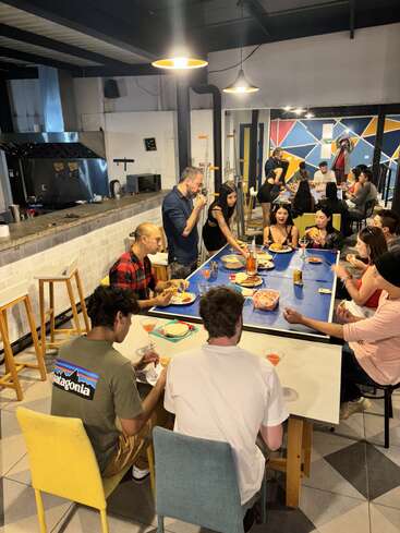 The image depicts a group of people gathered around a large table in a restaurant or cafeteria, enjoying food and drinks in a casual and relaxed atmosphere.