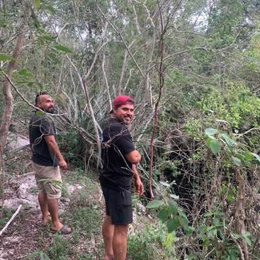 Two men stand in a wooded area, surrounded by trees and bushes, with one wearing a red cap and the other in shorts and a black t-shirt.
