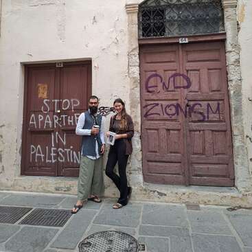 Two people stand in front of old wooden doors with political graffiti reading "STOP APARTHEID IN PALESTINE" and "END ZIONISM," on a worn stone street.