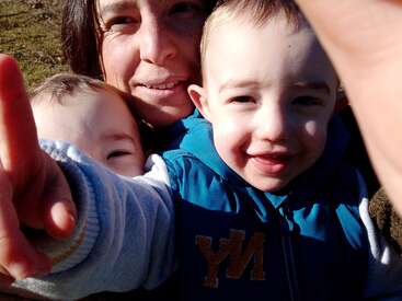 The image shows a woman holding two babies, with the woman\'s face blurred out, and the babies wearing blue jackets, smiling at the camera.