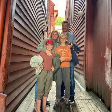 The image depicts a family of four posing for a photo on a wooden walkway between two buildings, with the family members standing close together.