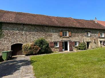 The image depicts a rustic stone building with a brown roof, featuring a green trash can, lush grass, and a serene blue sky.