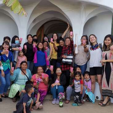 The image depicts a diverse group of approximately 20 people, including children and adults, gathered outside a white building with archways, holding pink cups and smiling.