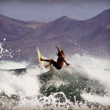 A man is surfing on a wave, with his surfboard in the air, wearing shorts, and with mountains in the background.