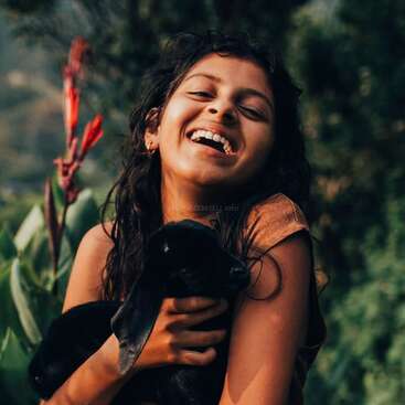 A young woman with long dark hair holds a black rabbit, smiling with eyes closed, set against a blurred green background with a red plant.