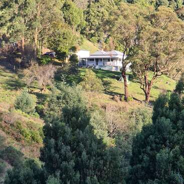 The image depicts a serene white house with a wraparound porch, nestled among trees on a hillside, surrounded by lush greenery and a vibrant natural landscape.
