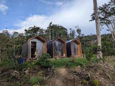 Three small, modern wooden cabins stand side by side on a lush, green hillside surrounded by trees, under a partly cloudy sky, evoking tranquility and nature.