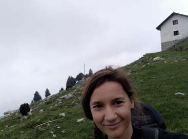 The image depicts a woman with brown hair and a backpack, smiling at the camera, set against a grassy hillside with rocks, trees, and a white building in the background.