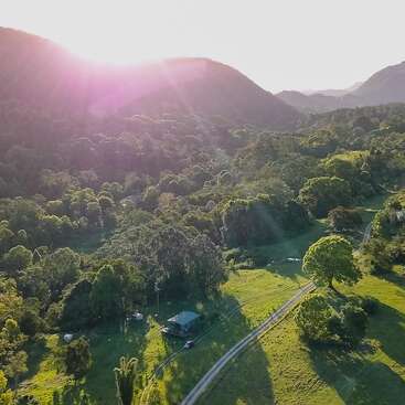 The image depicts a serene landscape featuring a winding road, a house, and lush greenery, with distant mountains and a radiant sun shining brightly in the sky.