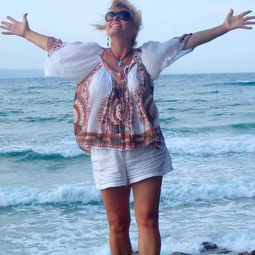 A woman stands on a rock at the beach, arms outstretched, wearing white shorts, a patterned blouse, and sunglasses, with the ocean and sky in the background.