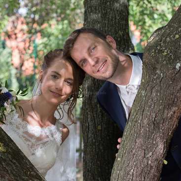 The image depicts a newlywed couple posing in front of a tree, with the bride on the left and the groom on the right, both smiling at the camera.