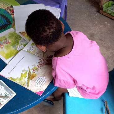 L'image montre un jeune enfant assis à une table bleue, lisant un livre aux illustrations et au texte colorés, vêtu d'une chemise rose et d'un short blanc.