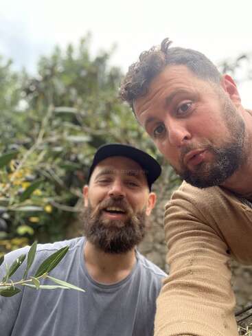 The image shows two men posing for a selfie in front of a blurred background of greenery and a stone wall. They both have beards.