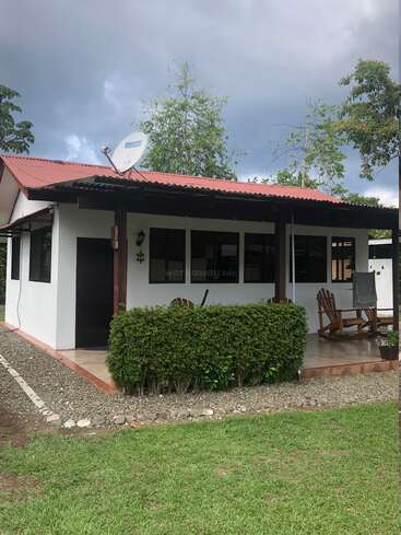 The image depicts a small, one-story white house with a red roof, featuring a covered porch and a satellite dish on the roof, set against a cloudy sky.