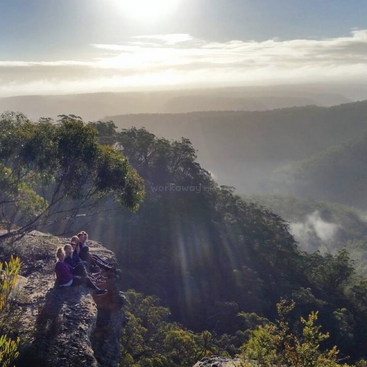 La imagen muestra a tres individuos sentados en un afloramiento rocoso, con vistas a un frondoso valle boscoso con colinas neblinosas a lo lejos, bañado por la luz del sol.