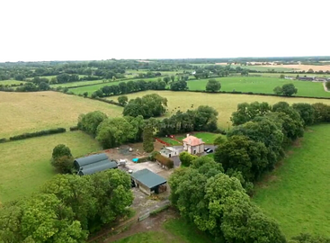 The image depicts a serene landscape featuring a large house with a sprawling yard, surrounded by trees and a fence, with a clear sky and a body of water in the background.