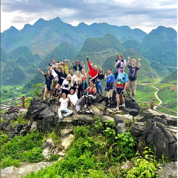 The image depicts a group of 18 people posing for a photo on a rocky outcrop, set against a stunning backdrop of lush green mountains and a winding road.